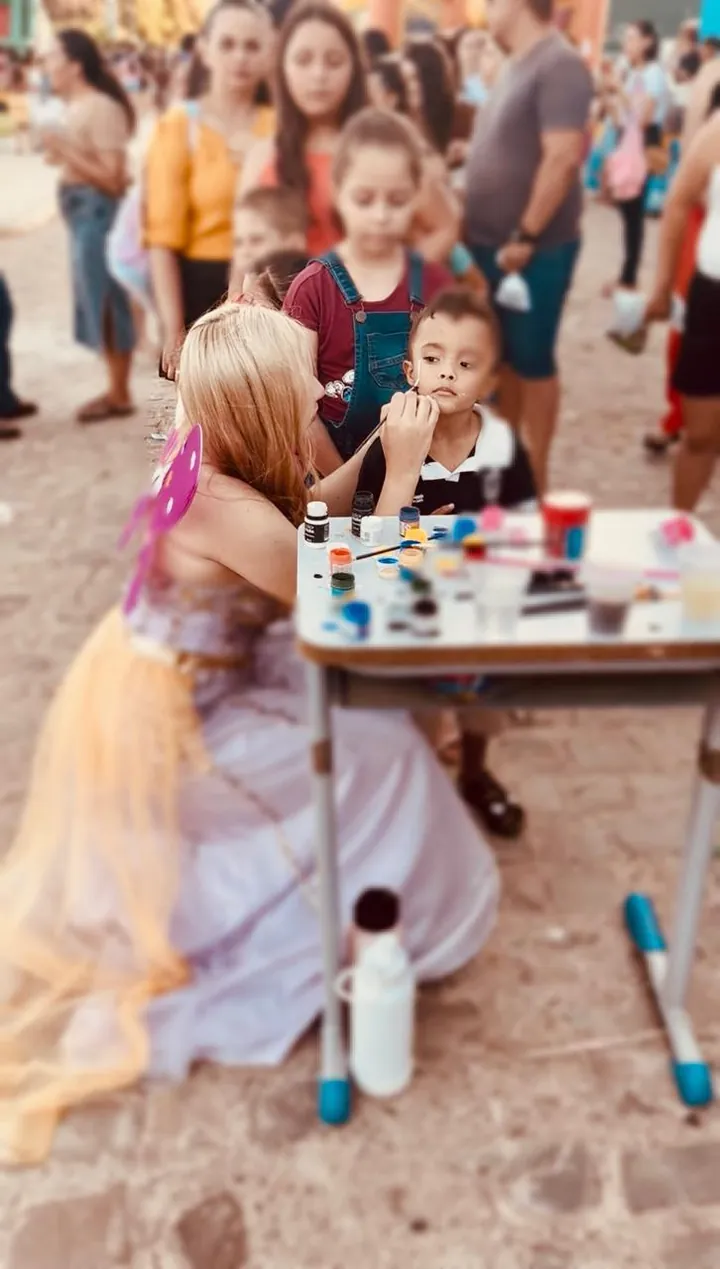 Lanna Campos fantasiada, fazendo pintura facial em uma criança em uma festa ao ar livre.
